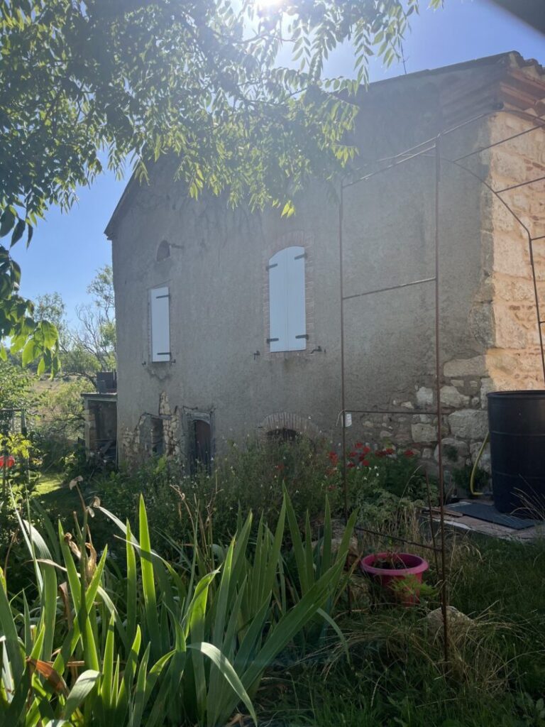 Ancienne maison en rénovation énergétique, façade avec volets bleus, jardin luxuriant.